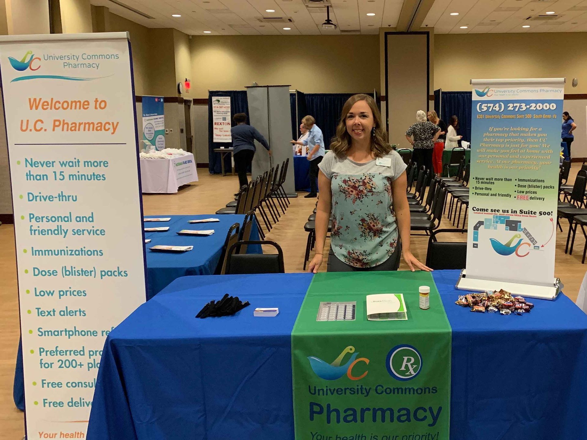 UC Pharmacy outreach booth at a health fair, with a smiling staff member behind a blue table display.