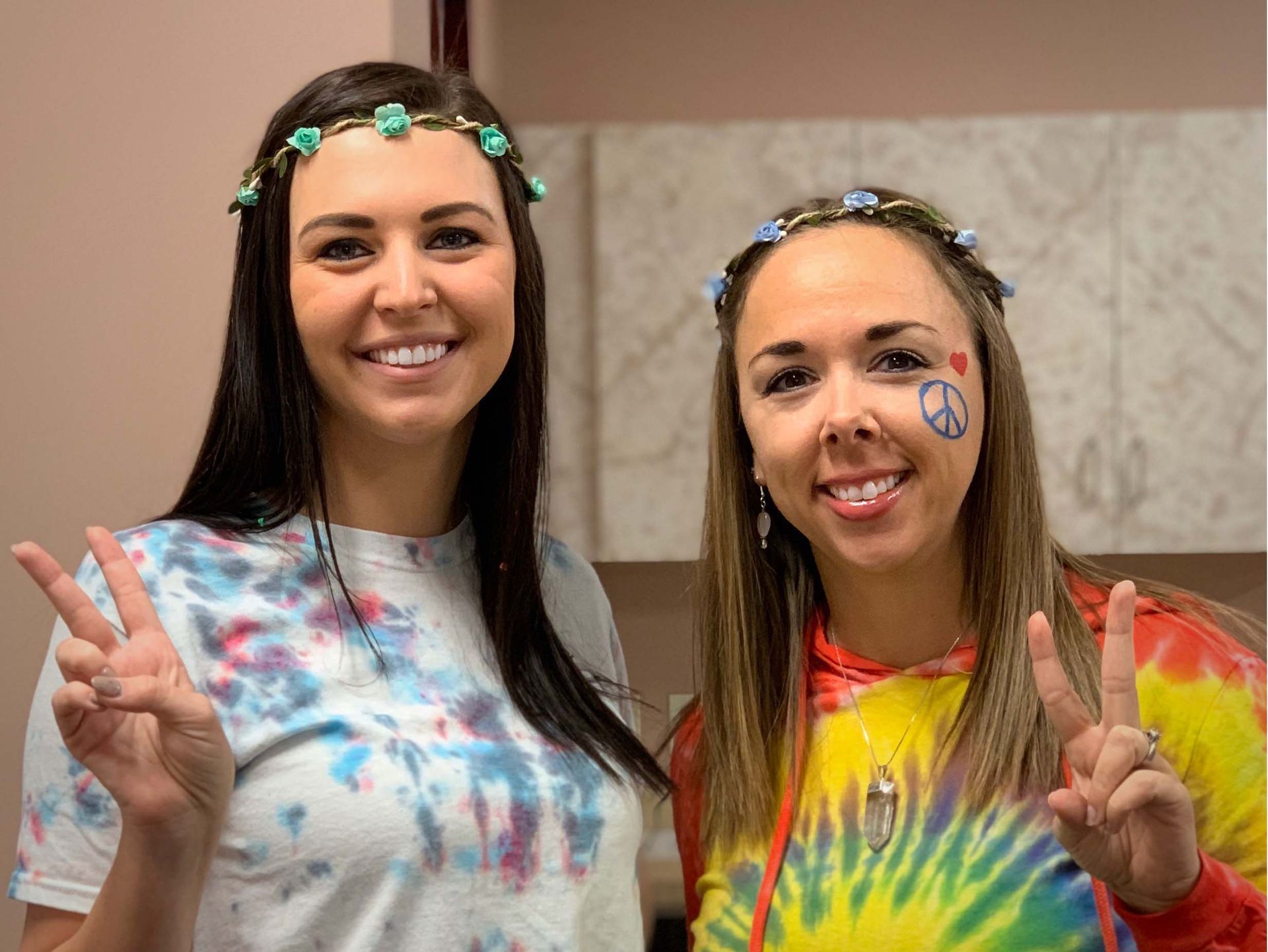 Two smiling women in colorful tie-dye shirts pose with peace signs indoors.