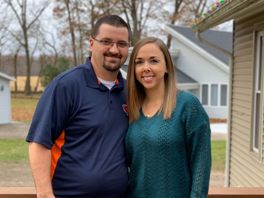 Smiling couple standing outdoors by a house, with trees and a cloudy sky in the background.