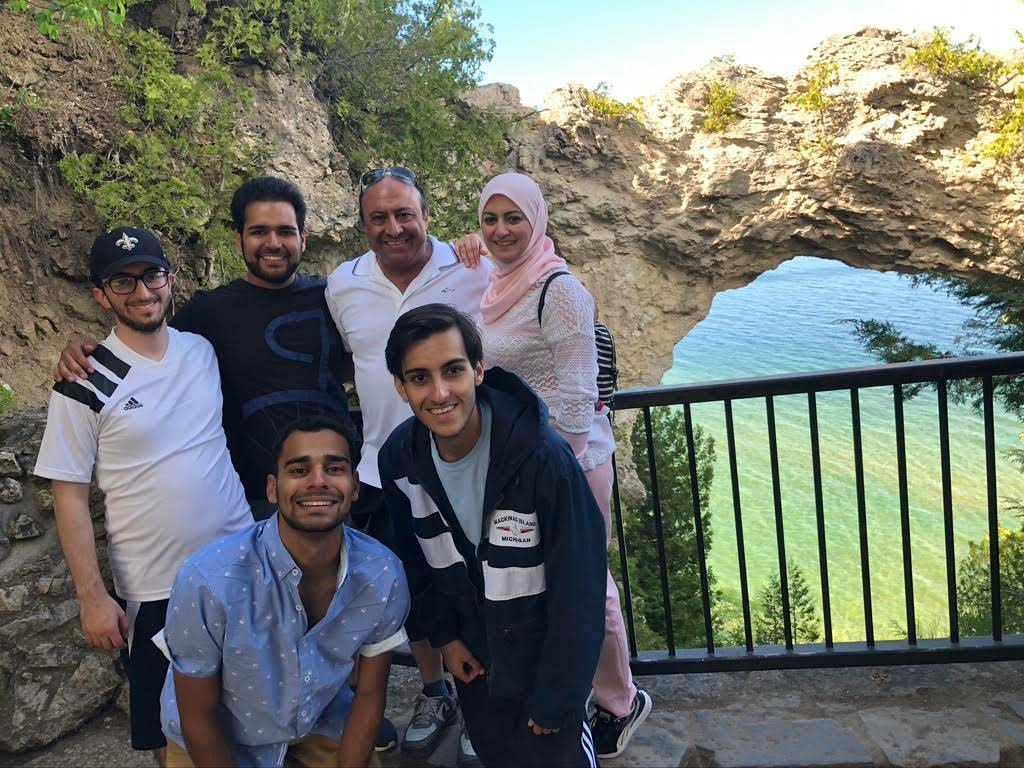 Group of six smiling people posing by a metal railing with a scenic blue-green lake and rocky hillside behind them