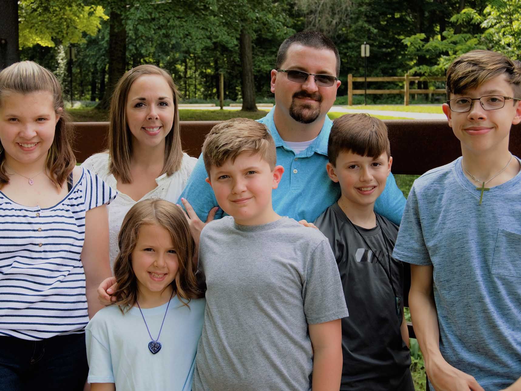 Group of six smiling people posing outdoors in front of trees and a wooden fence