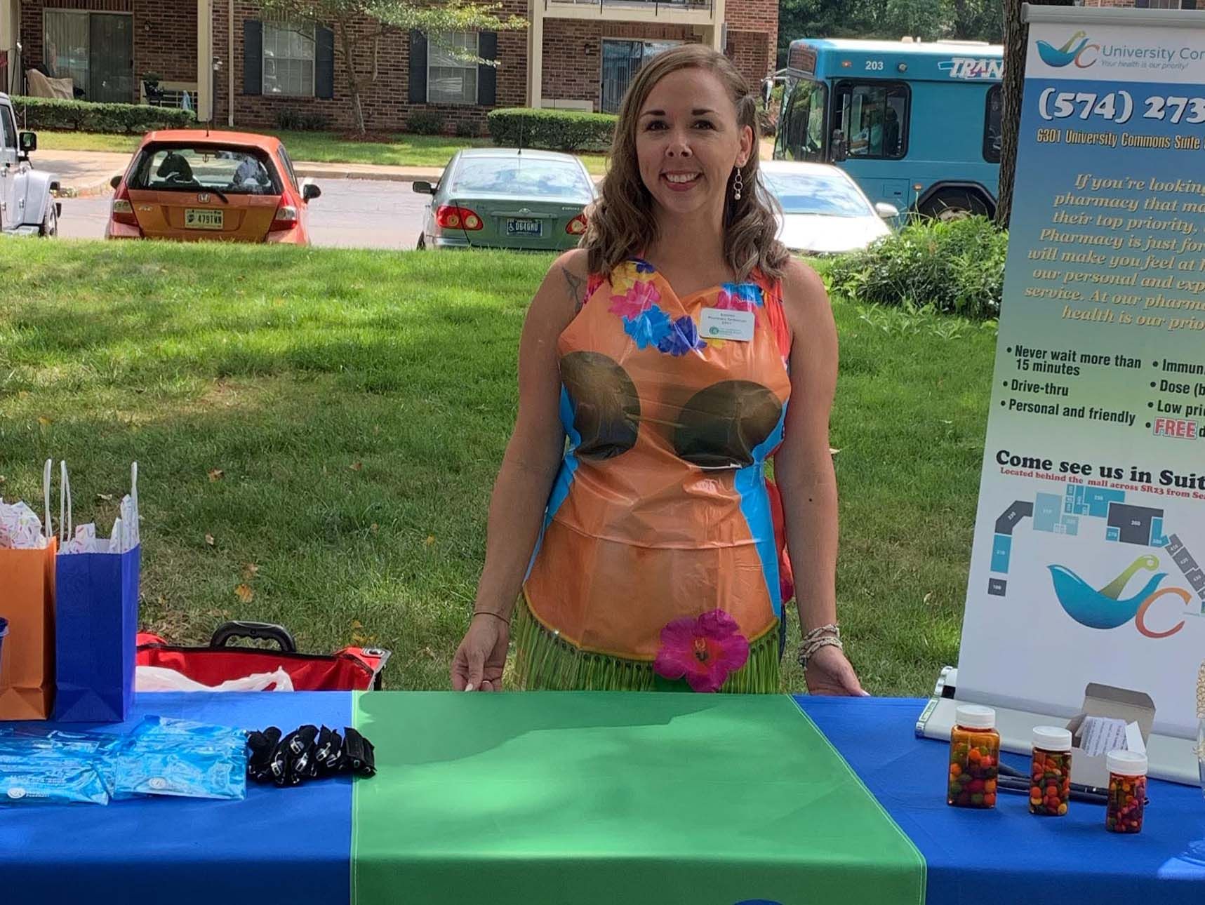 Woman in an orange, blue, and pink costume stands behind a green table at an outdoor event.