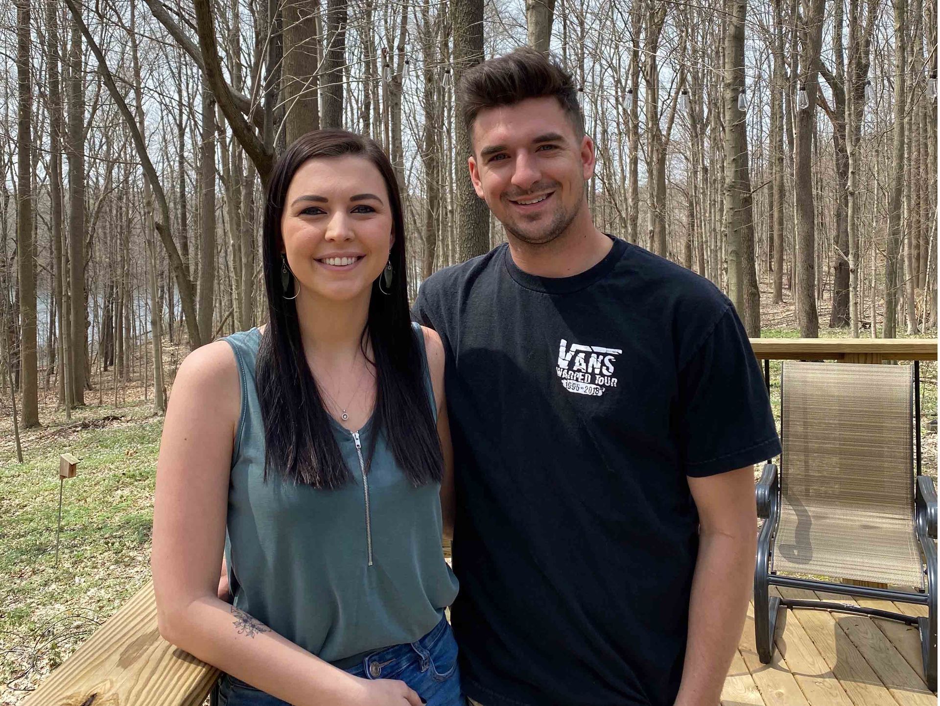 Two people standing outdoors in a wooded area, smiling at the camera.
