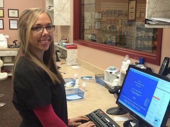 Woman in black scrubs stands at a clinic counter beside a computer, smiling at the camera.