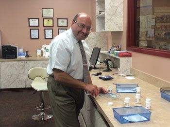 Man standing at a reception desk, smiling in an office with chairs, frames, and blue trays on the counter.