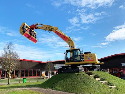 Yellow excavator on a grassy mound beside a building, lifting a red mower attachment under a blue sky