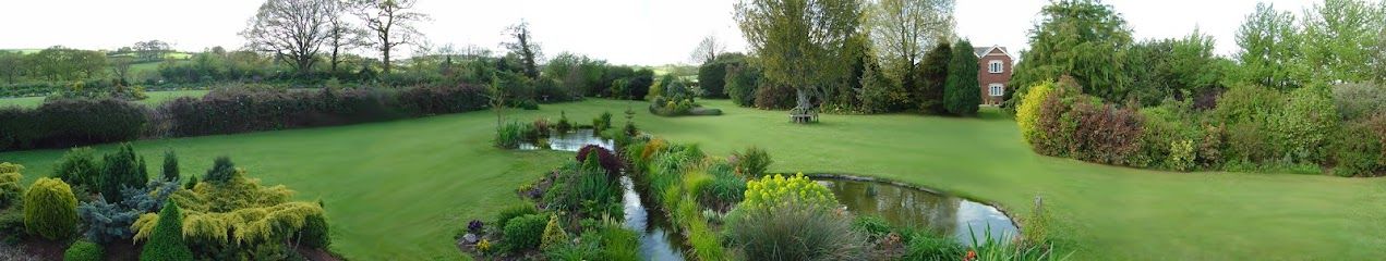 Panoramic garden with lush green lawn, flower beds, shrubs, and trees under a cloudy sky