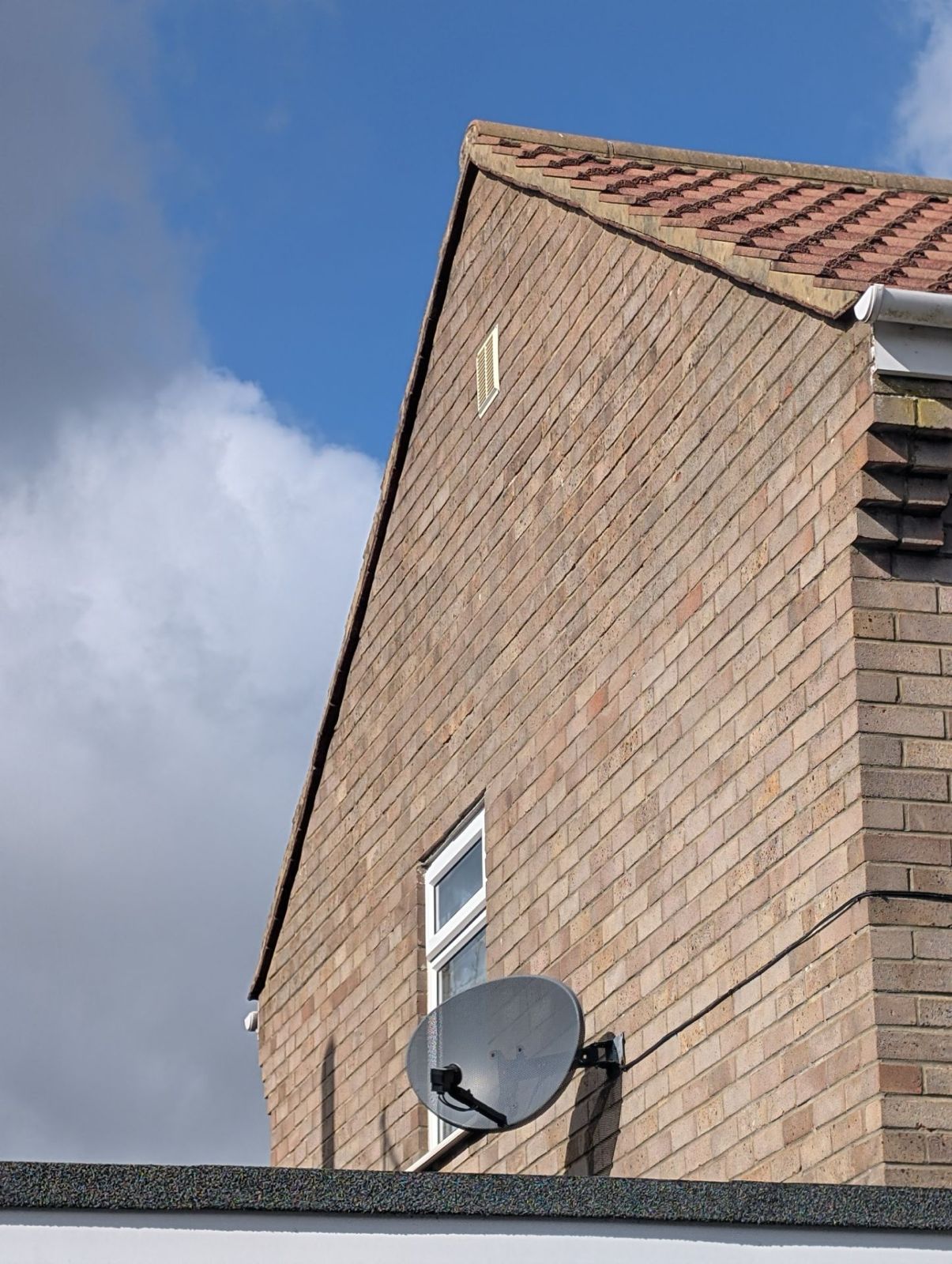 Brick house gable with tiled roof, small window, and satellite dish against a blue sky