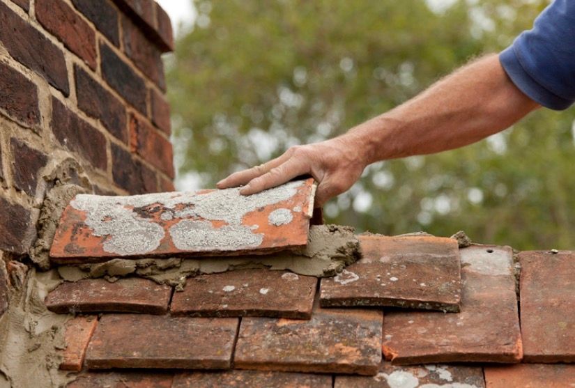 Hand placing a brick onto a partially repaired brick wall beside a chimney