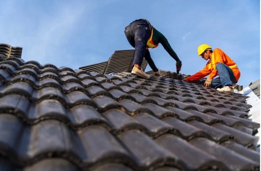 Two workers repairing a tiled roof, one in black and one in orange safety gear.