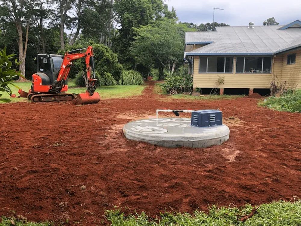 An Excavator is Working on a Septic Tank in Front of a House — Bevan Plumbing in Bangalow, NSW