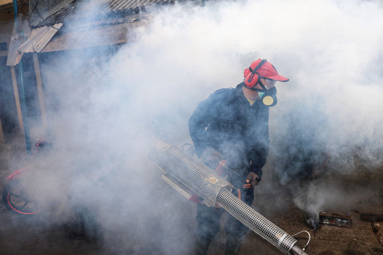 Person in protective gear sprays insecticide, surrounded by smoke.