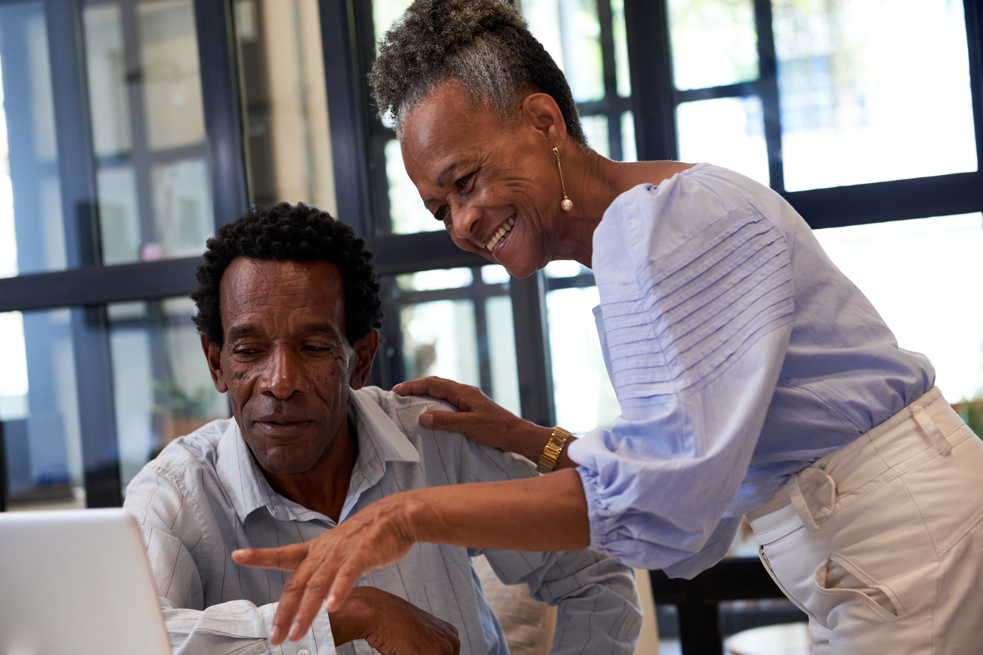 Older Black couple looking at a laptop. Woman points and smiles; man looks focused. Indoors.