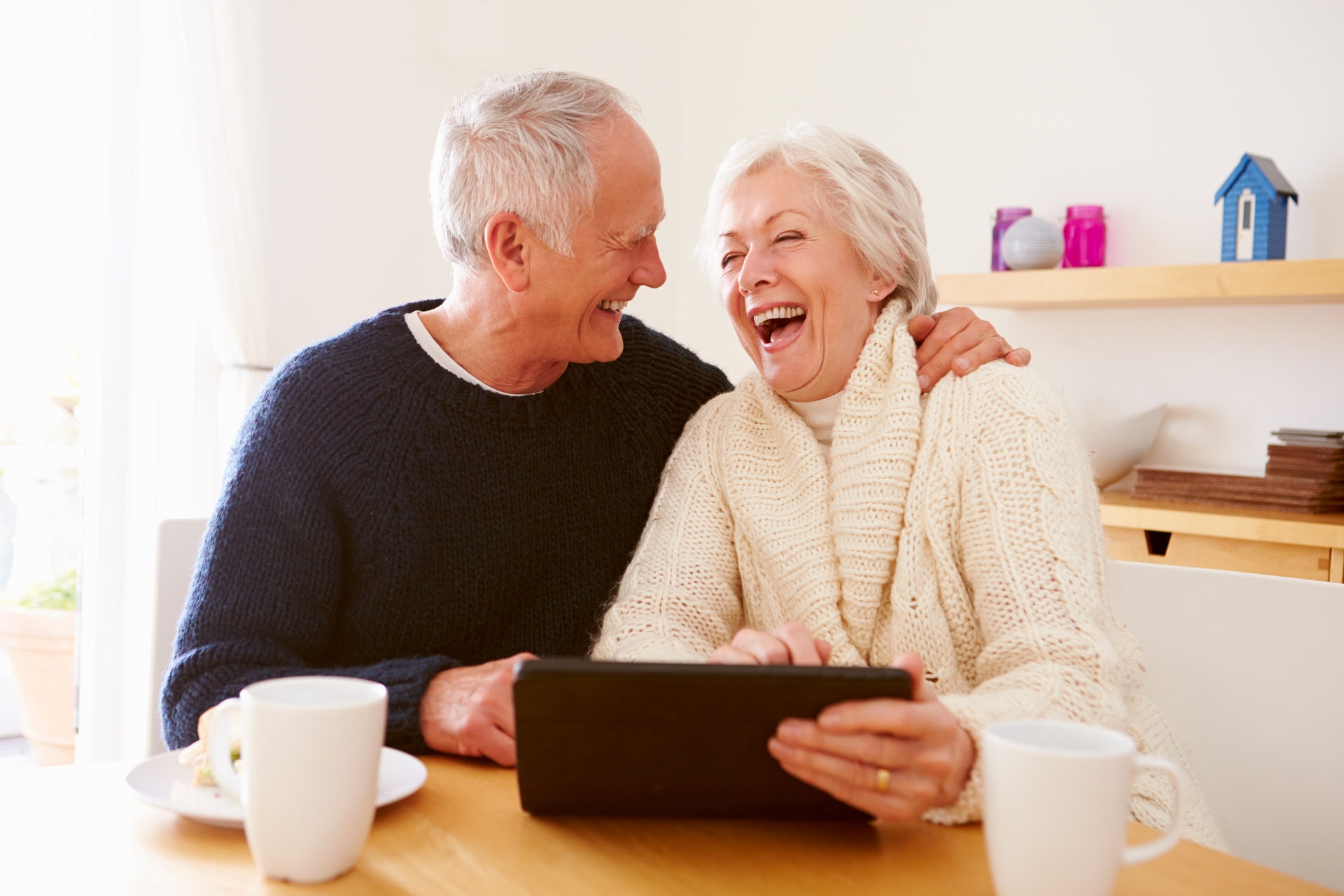 An elderly couple is sitting at a table using a tablet computer.