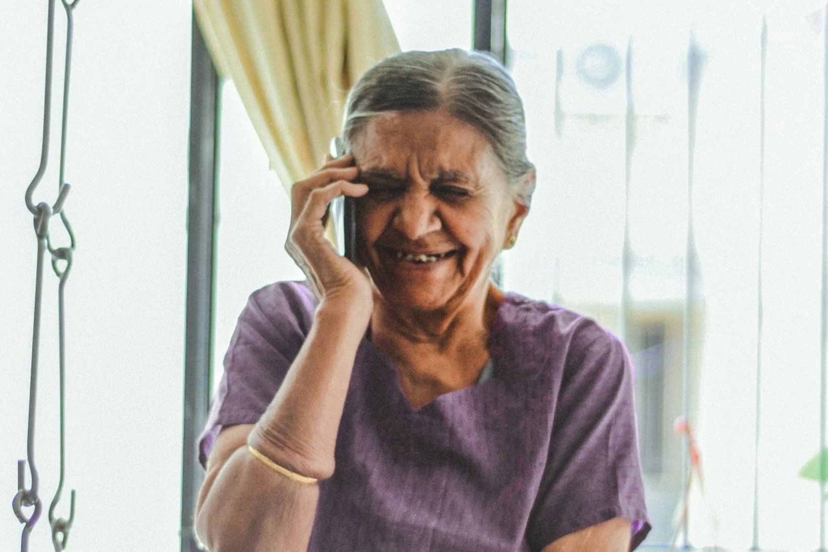 Elderly woman smiles while holding phone to her ear indoors, wearing purple shirt.