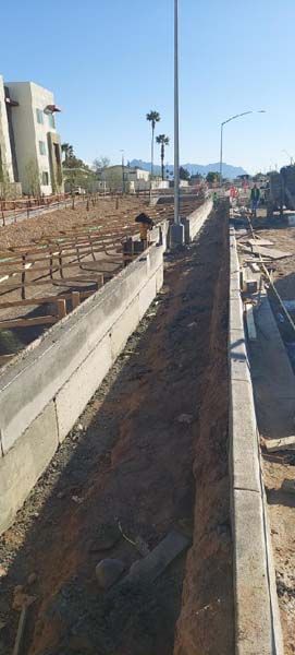 Construction site with a long, narrow trench between concrete walls. A building and palm trees are in the distance.