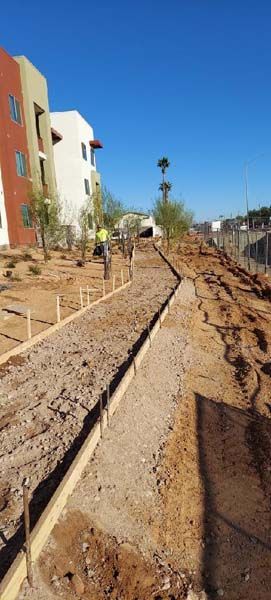 Construction site with dirt pathway framed by wood. Buildings and blue sky in the background.