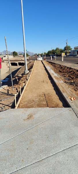 Construction site: sidewalk being built along a road with a dirt median. Clear blue sky.