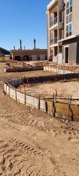 Construction site with wooden concrete forms. Building in background, tire tracks in the dirt. Blue sky.