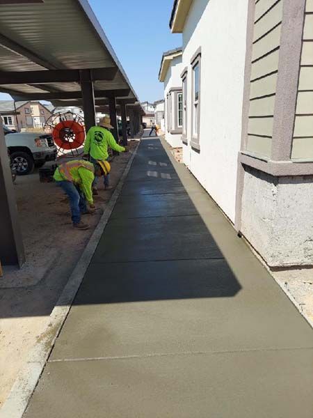 Workers pouring and smoothing wet concrete sidewalk next to a building with windows and a carport.