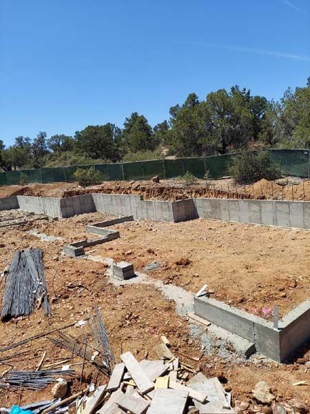 Construction site with concrete foundation walls, rebar, and dirt. Sunny day, trees in background.