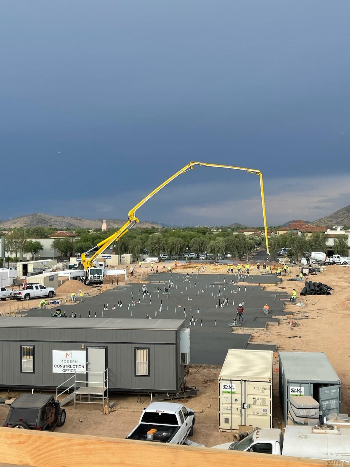 Construction site with concrete pump pouring concrete onto a large foundation; workers in the background under a stormy sky.