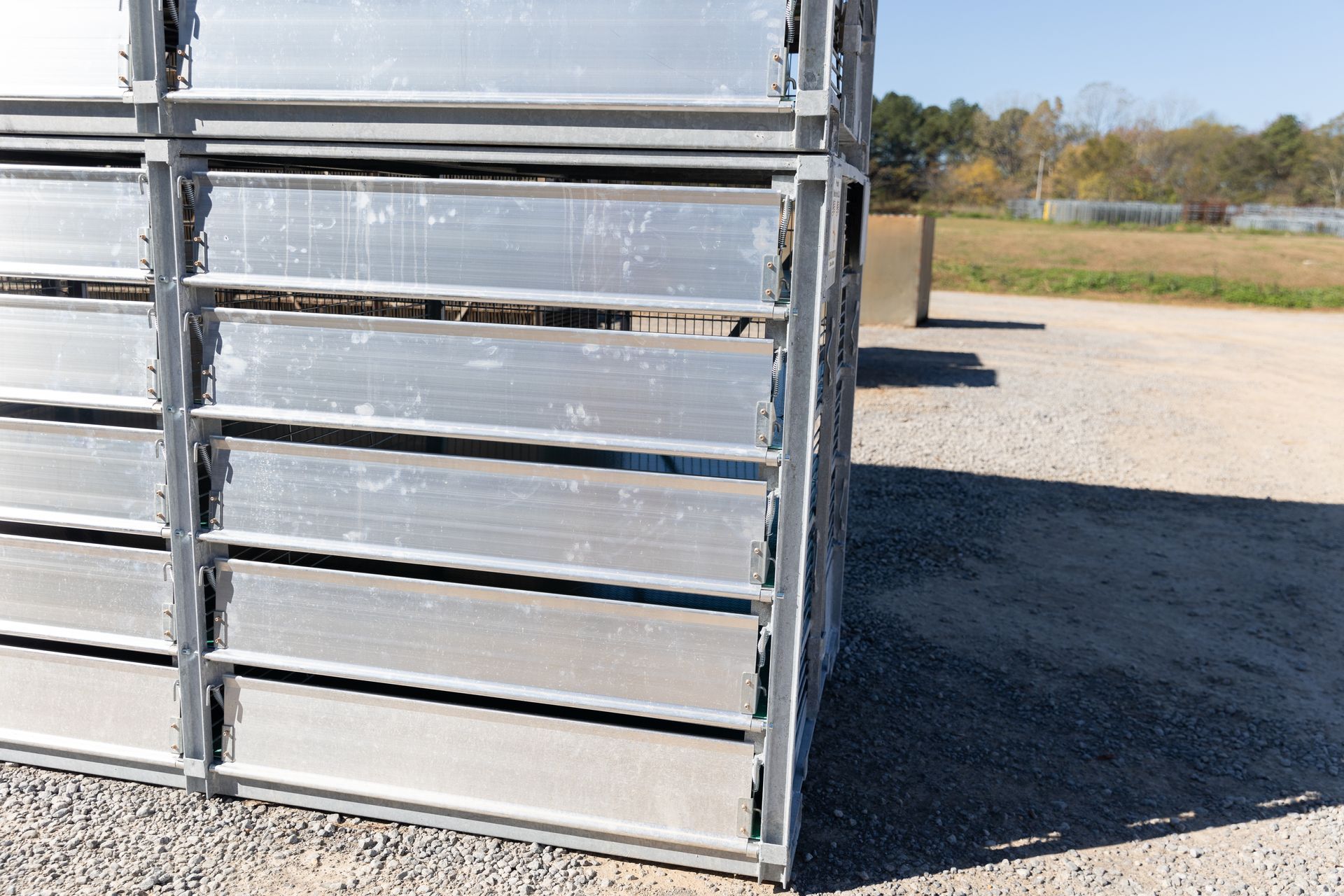 Metal panels stacked outdoors, on gravel, with a field and trees in the background.