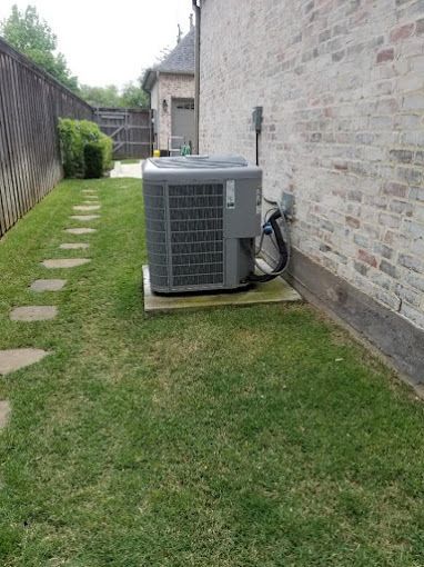 An air conditioner is sitting in the grass next to a brick wall.