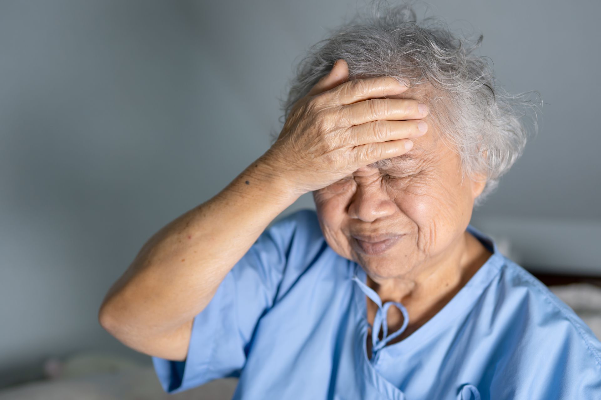 Elderly woman with gray hair, holding her hand to her forehead, appearing to have a headache. Wearing a blue hospital gown.