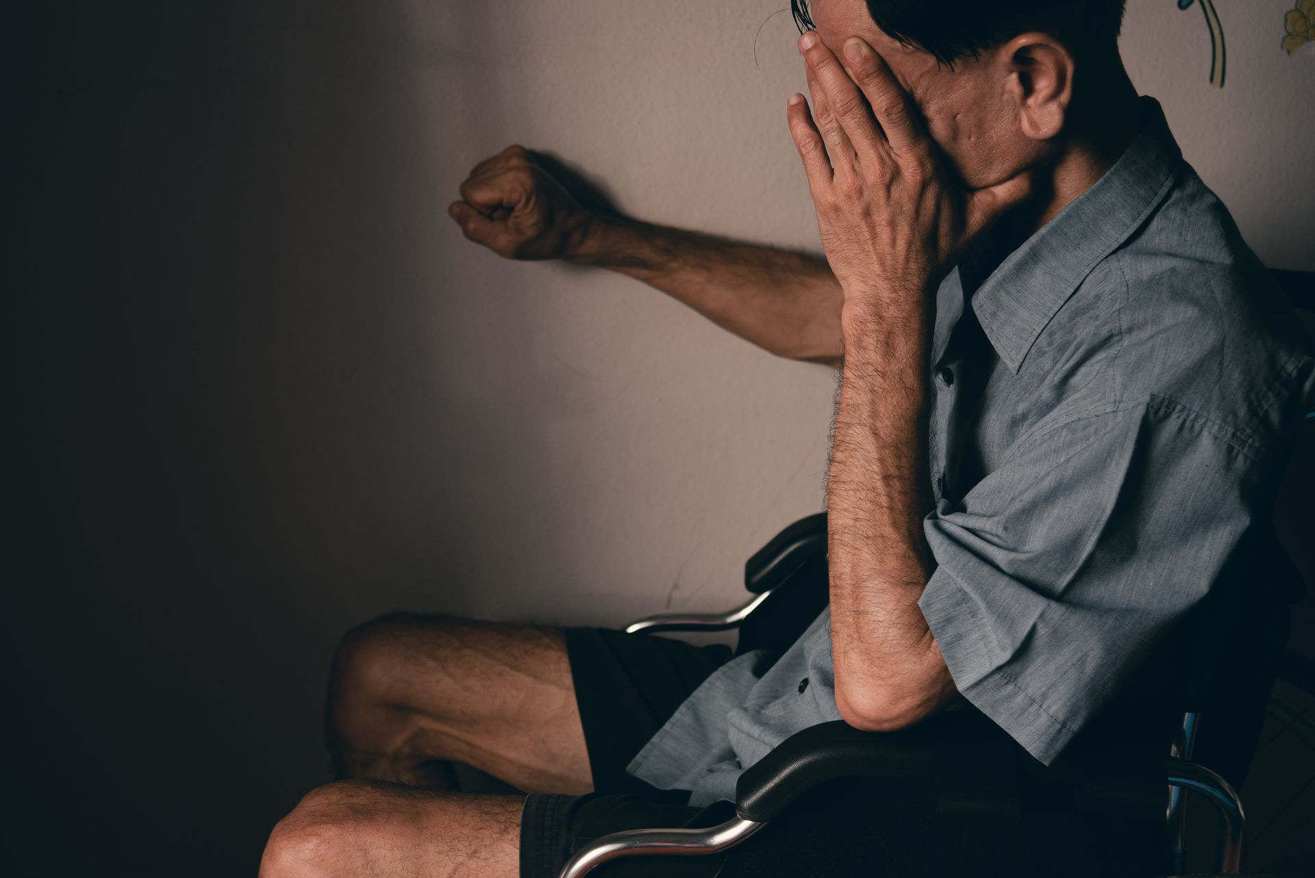 Man in wheelchair, hand covering face, fist raised towards wall. Lonely.
