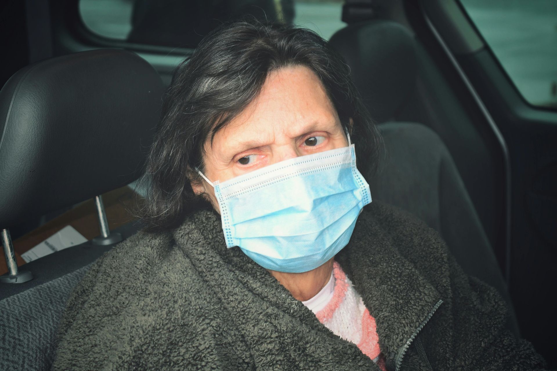 Woman in a surgical mask inside a vehicle, looking towards the viewer with a concerned expression.