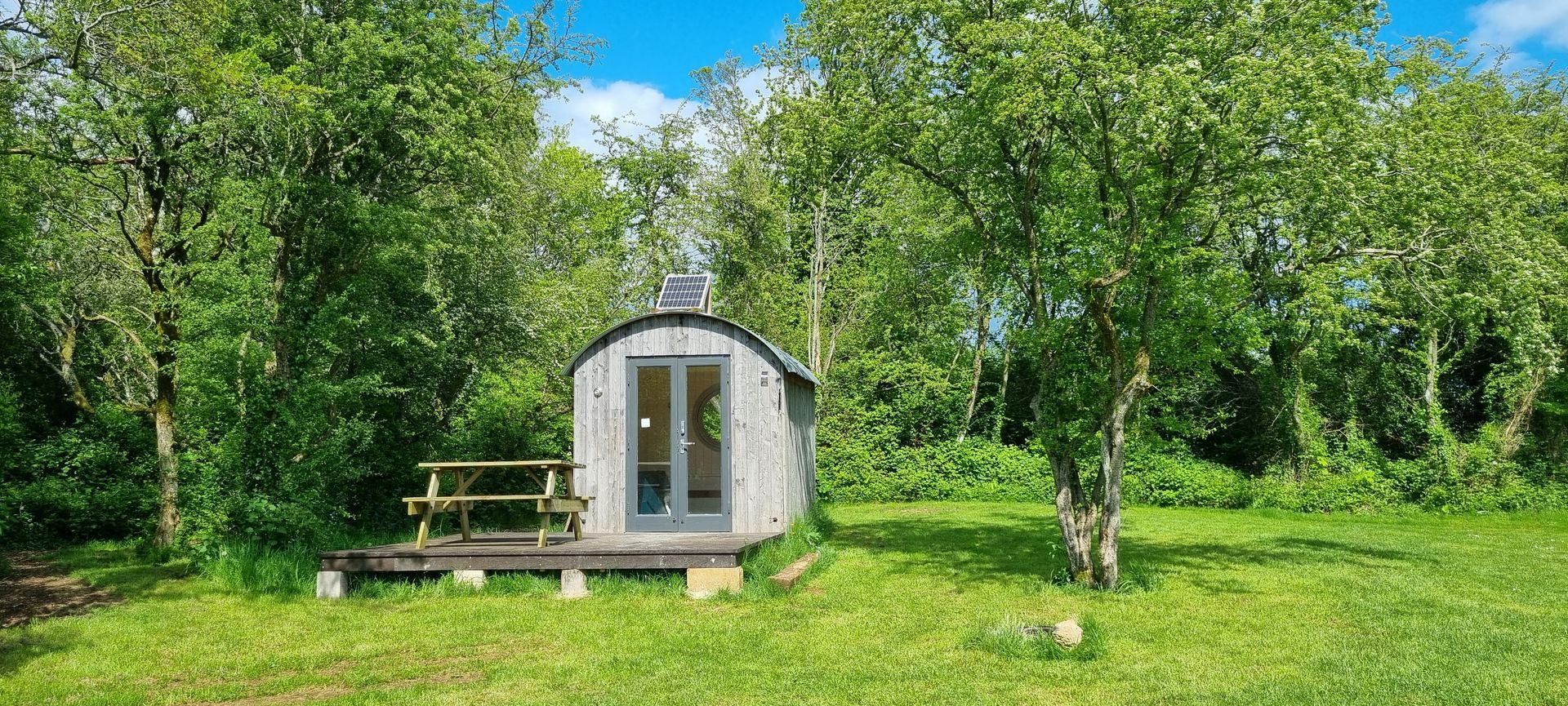 A small house with a picnic table in the middle of a field surrounded by trees.