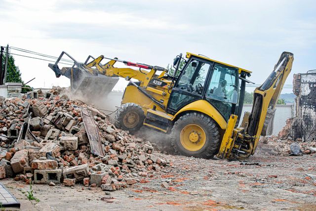 Yellow backhoe loader clearing rubble at a demolition site, kicking up dust.