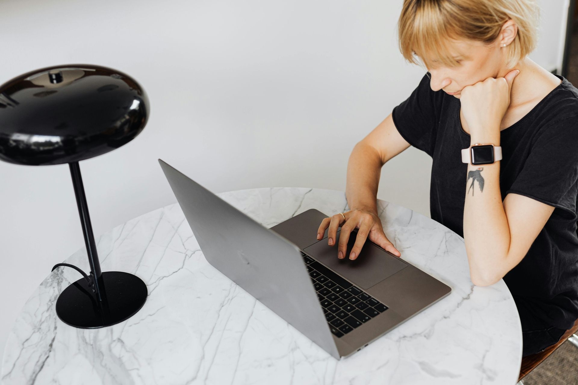 Woman with blonde hair using a laptop, resting her chin on her hand, at a white table with a black lamp.