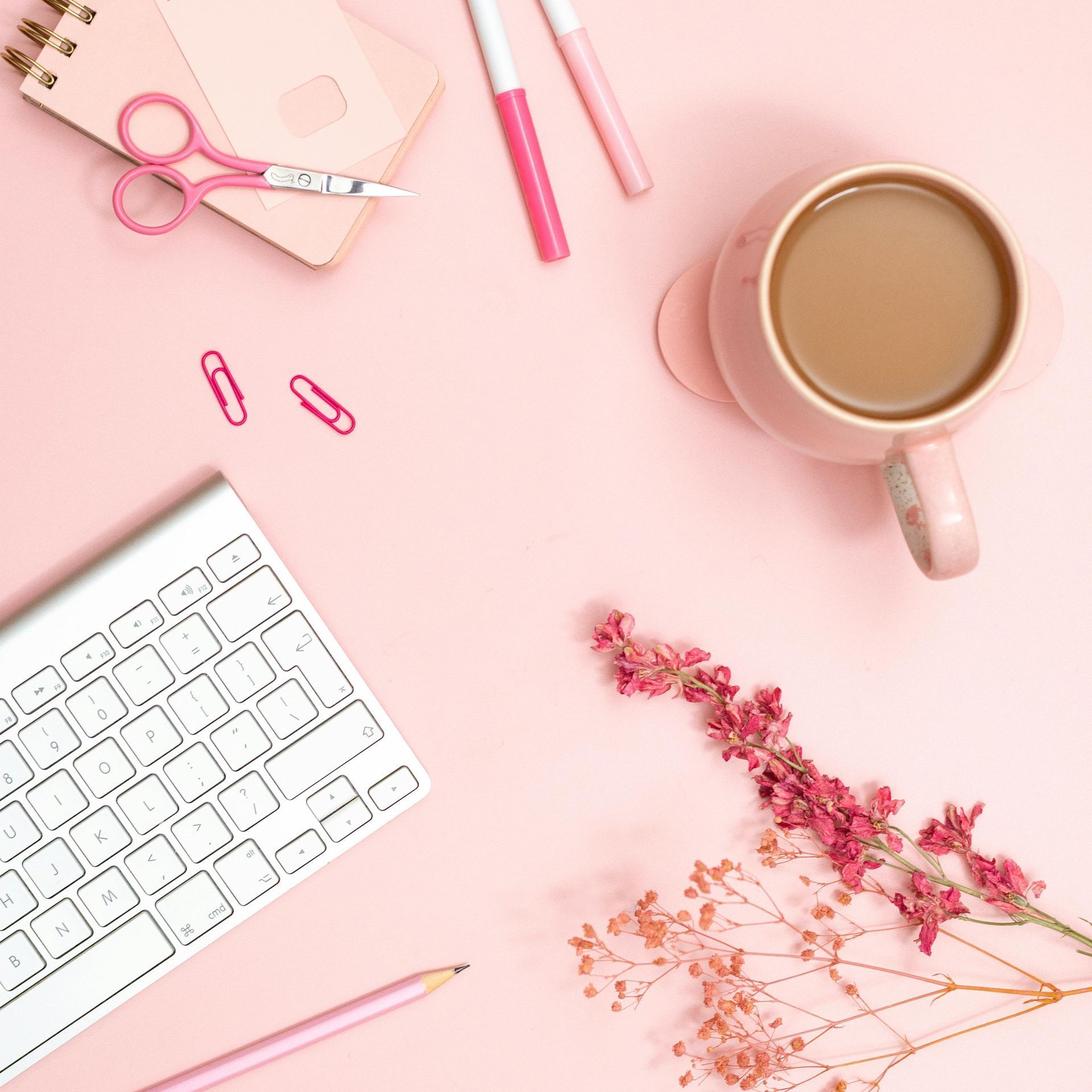 Pink desk with keyboard, coffee cup, stationery, and flowers.