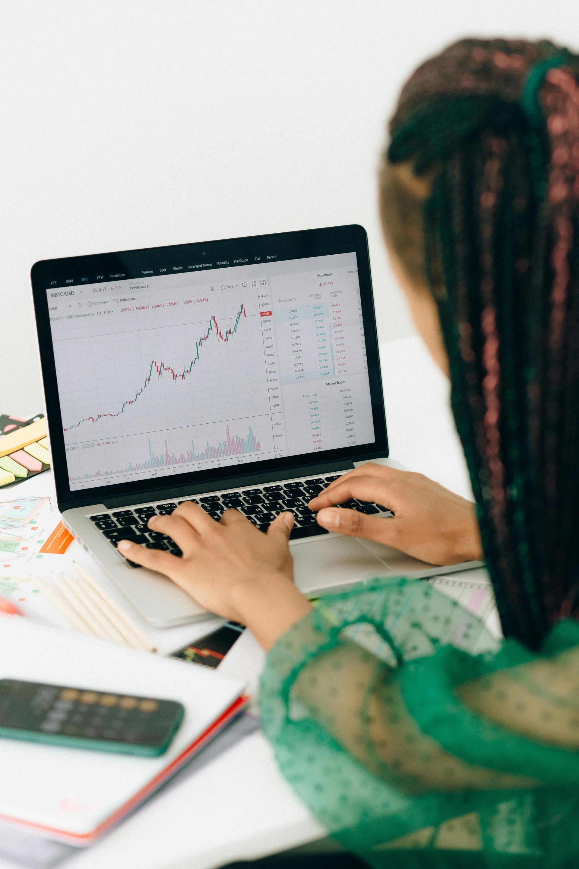Woman analyzing stock charts on a laptop, hands on the keyboard, wearing green, with braided hair.