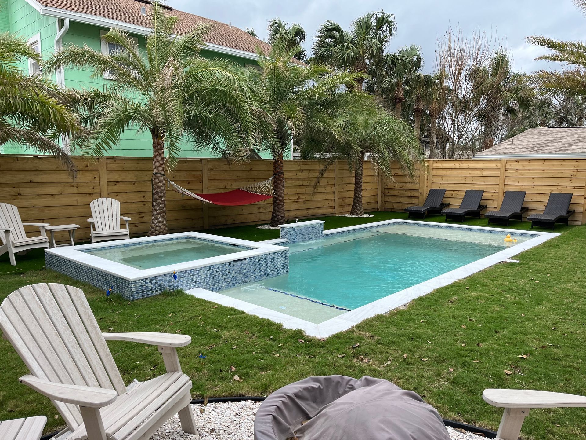 A large swimming pool in a backyard with chairs and a fire pit.