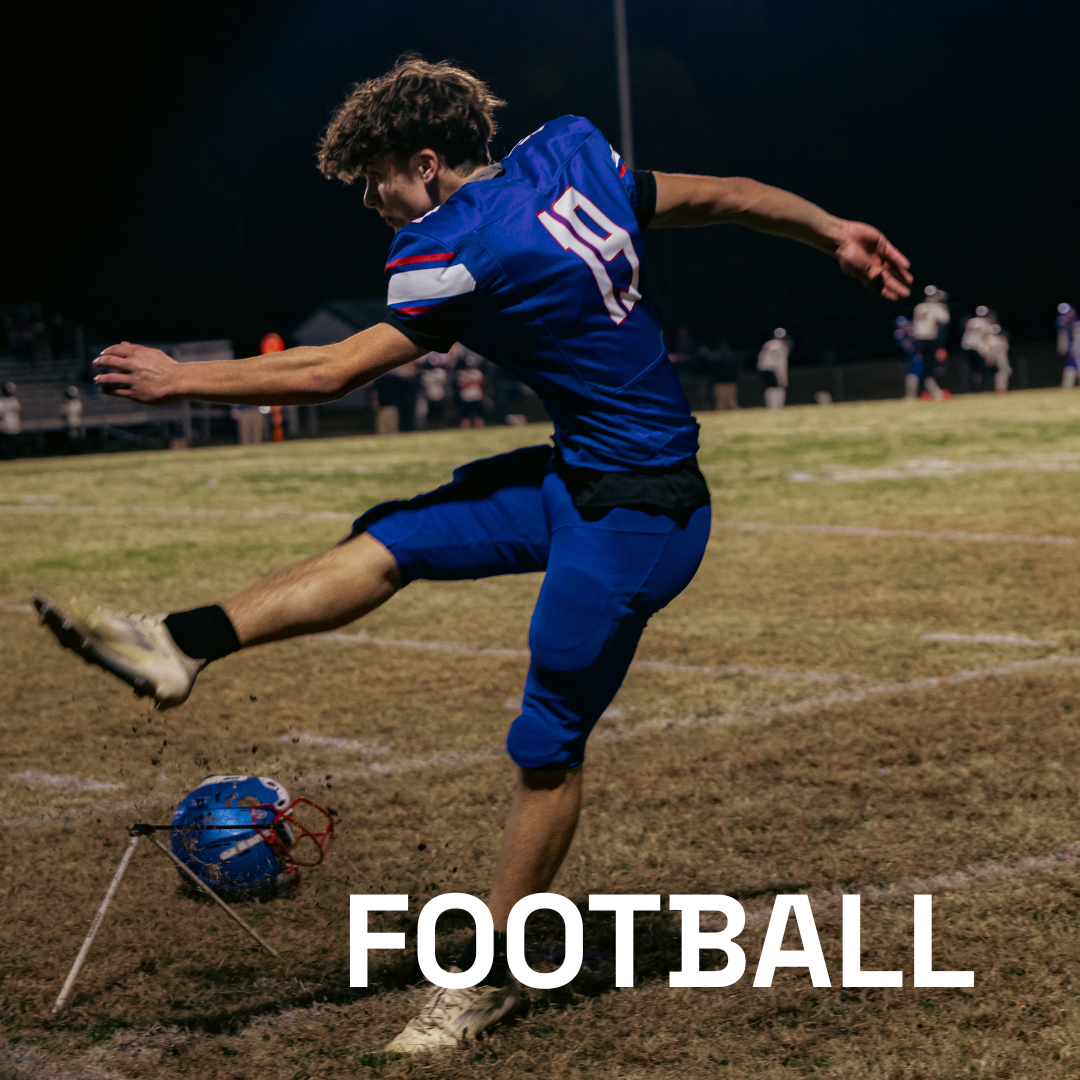 A player in a blue uniform kicks a football off a tee on a field during a night game.