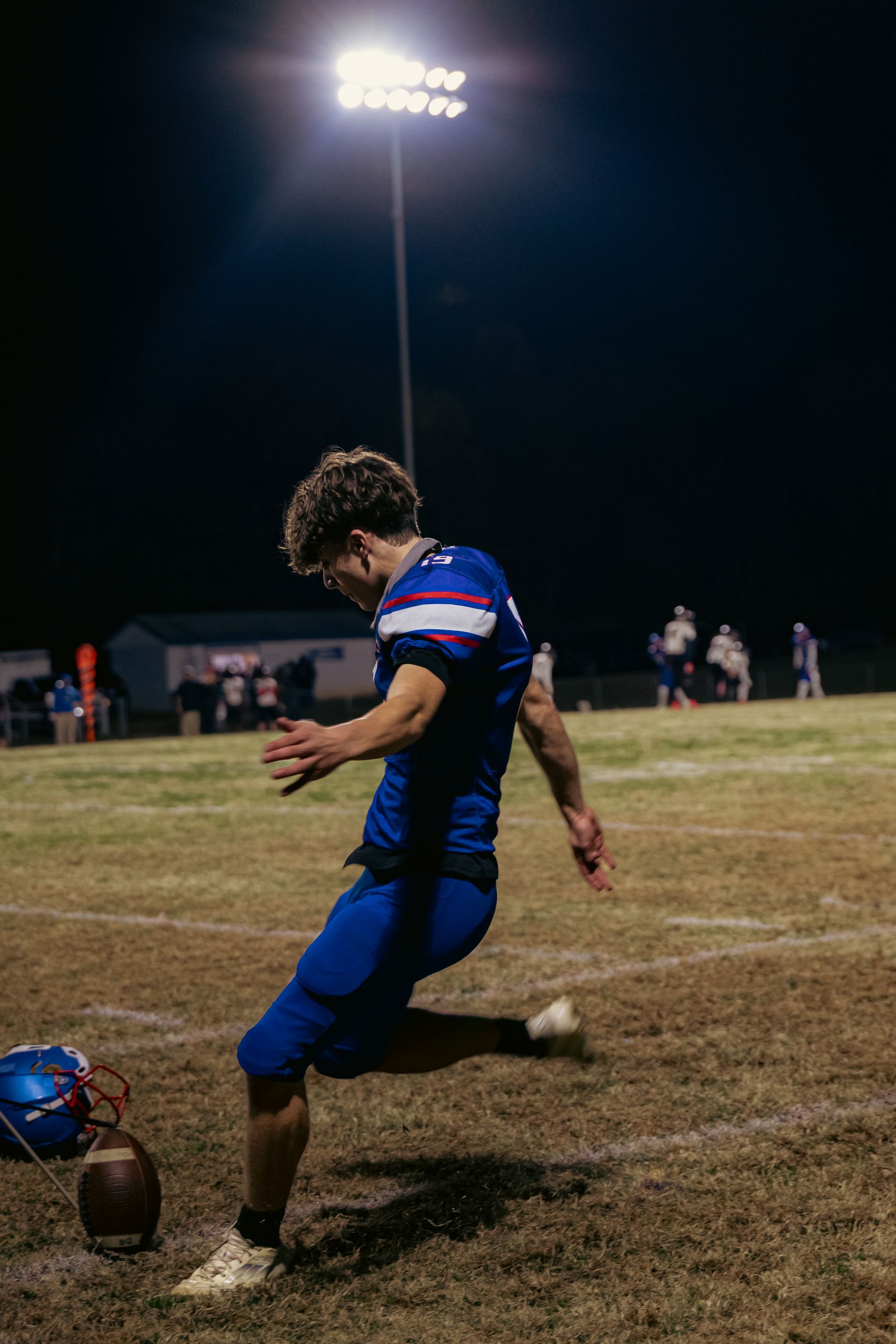 A football player in a blue uniform kicks a football on a field under bright stadium lights at night.