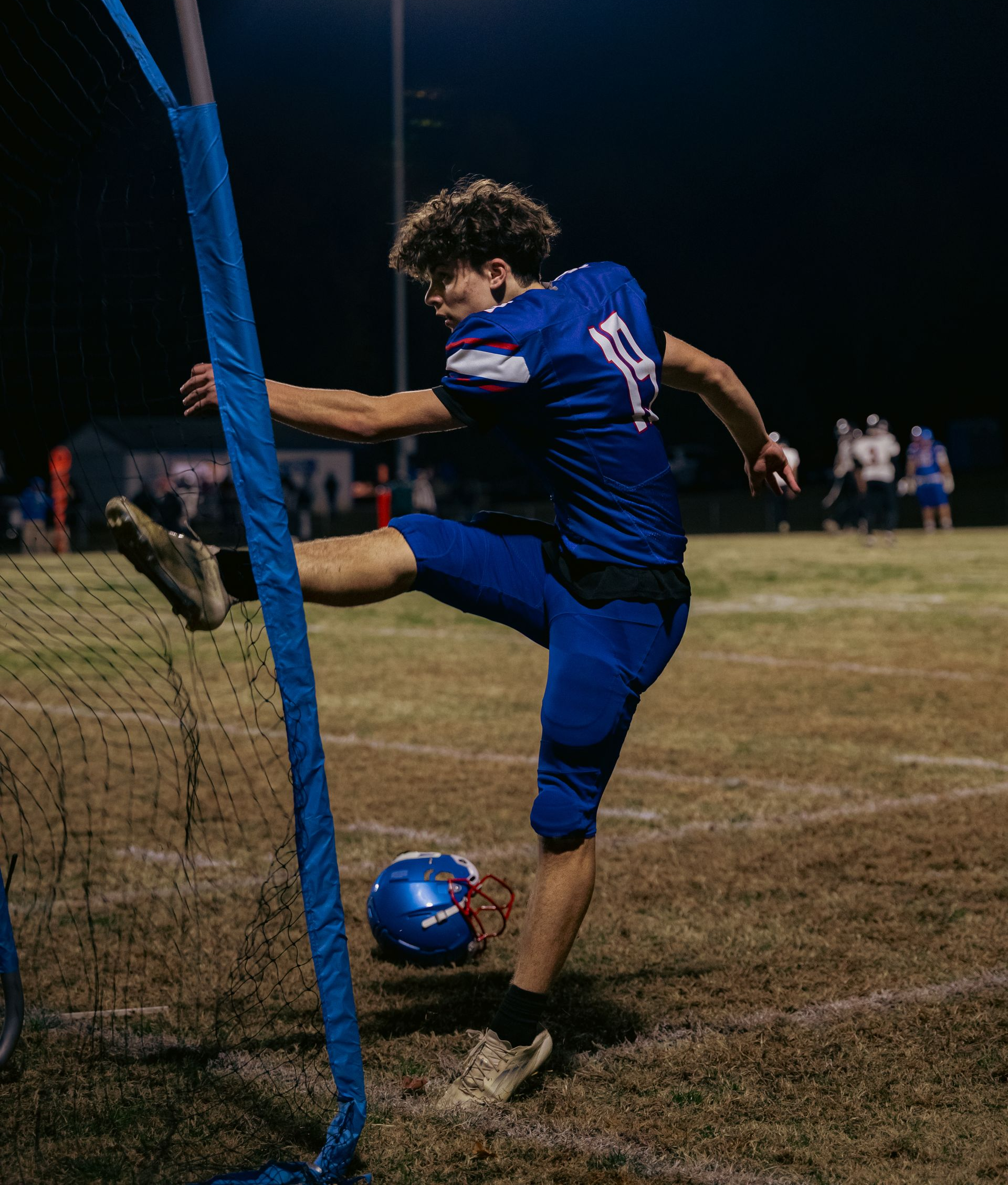 A football player in a blue uniform kicks a ball near a field goal post during a night game.