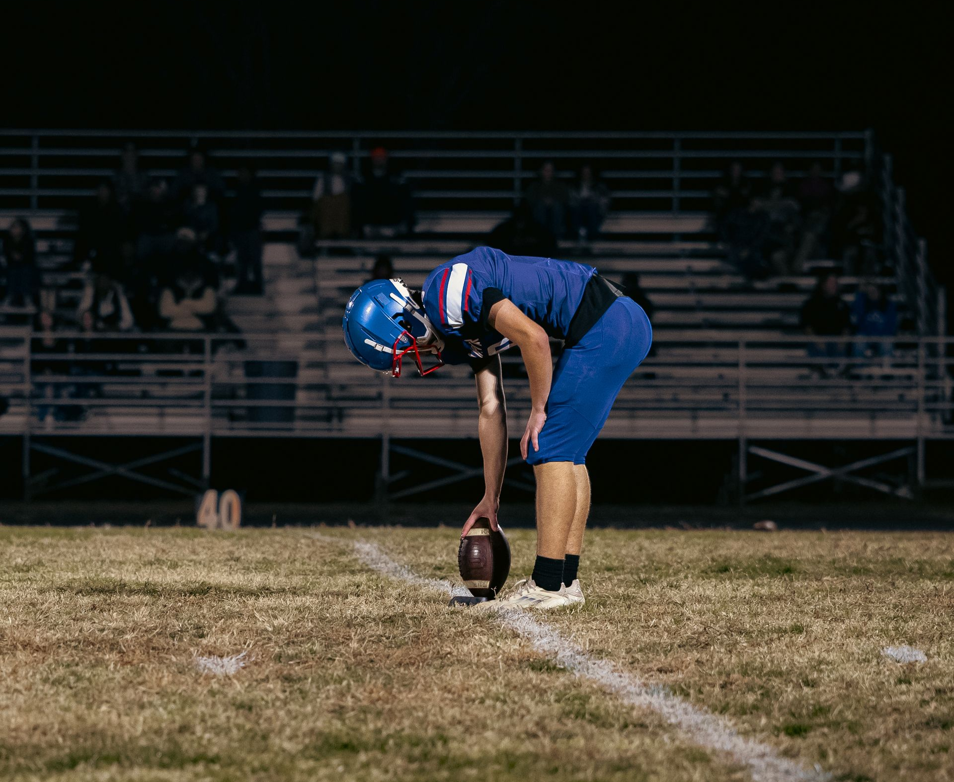 A football player in a blue uniform bends over to place a football on the field at night in front of empty stadium bleachers.