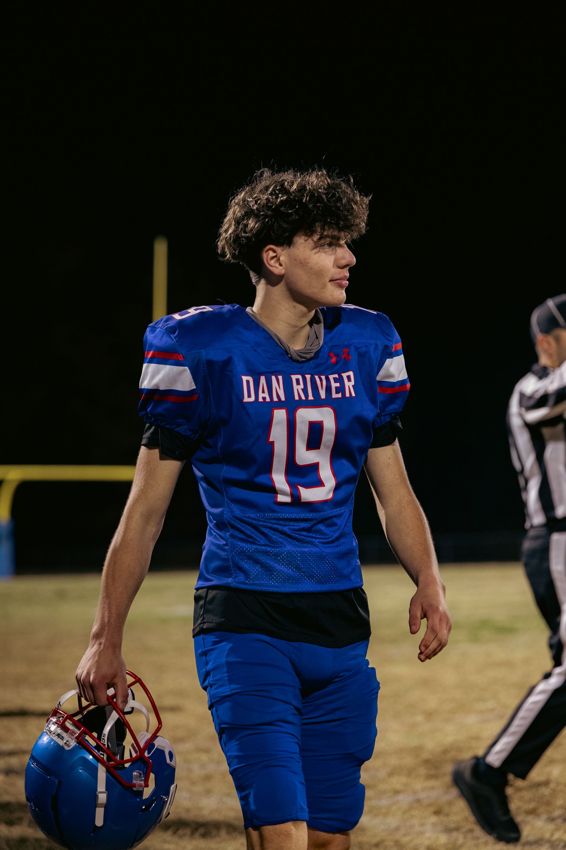 A football player in a blue Dan River jersey with the number 19 walks across a field at night, holding a helmet.