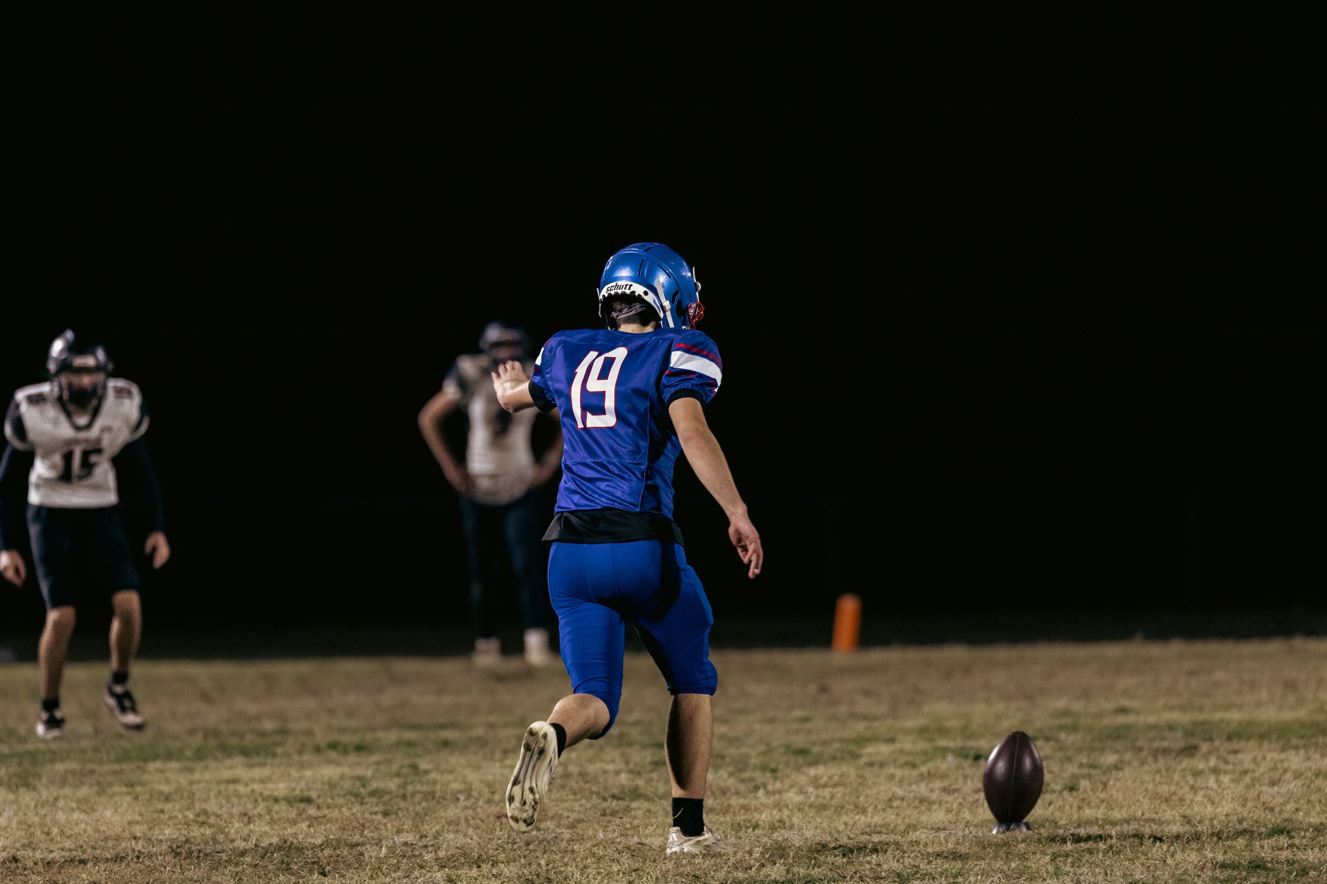 A football player in a blue jersey number 19 prepares to kick a football off a tee on a field at night.