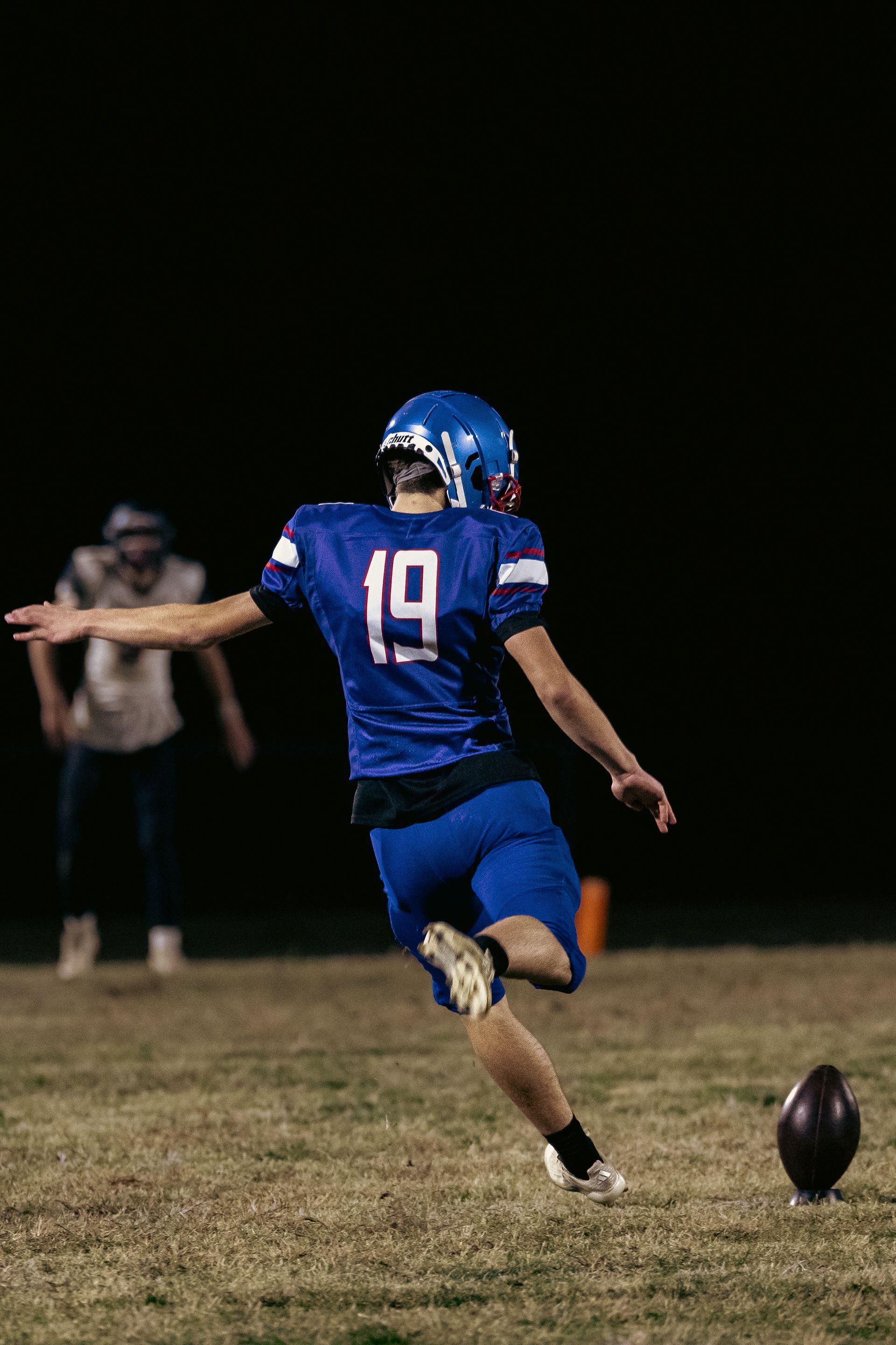 A football player in a blue uniform kicks a football on a field at night.