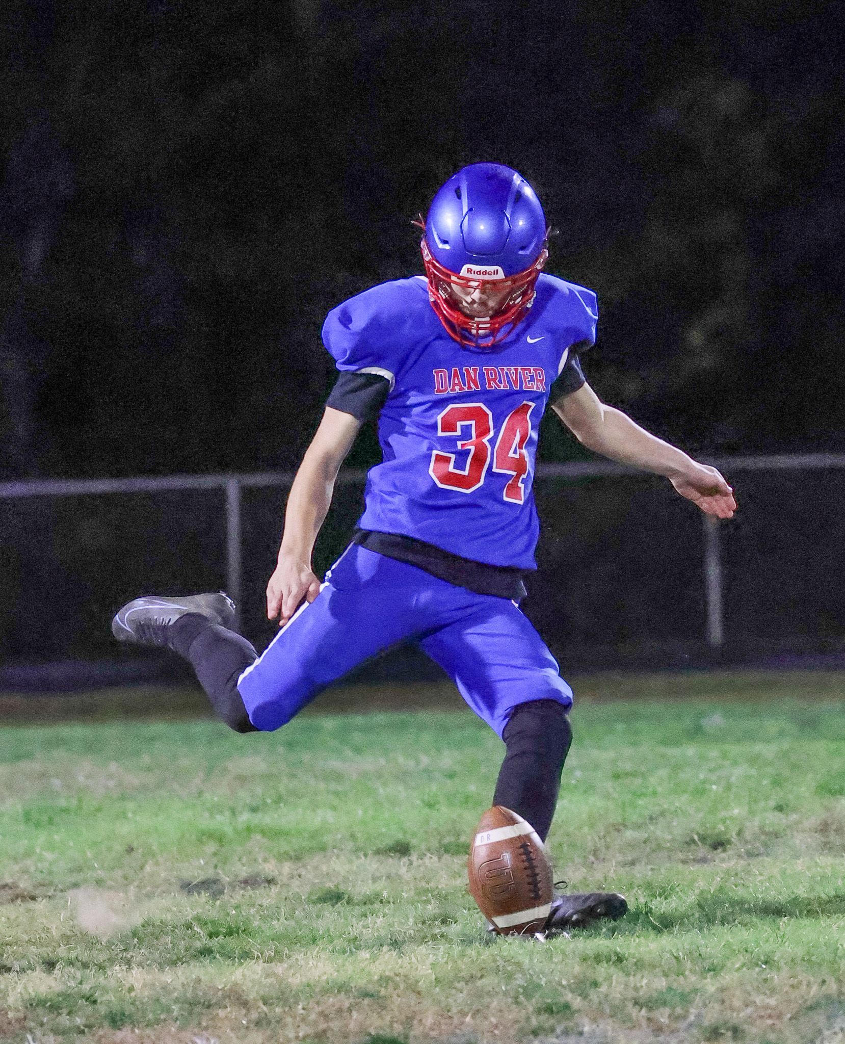 A football player wearing a blue uniform with the number 34 kicks a football on a grass field at night.