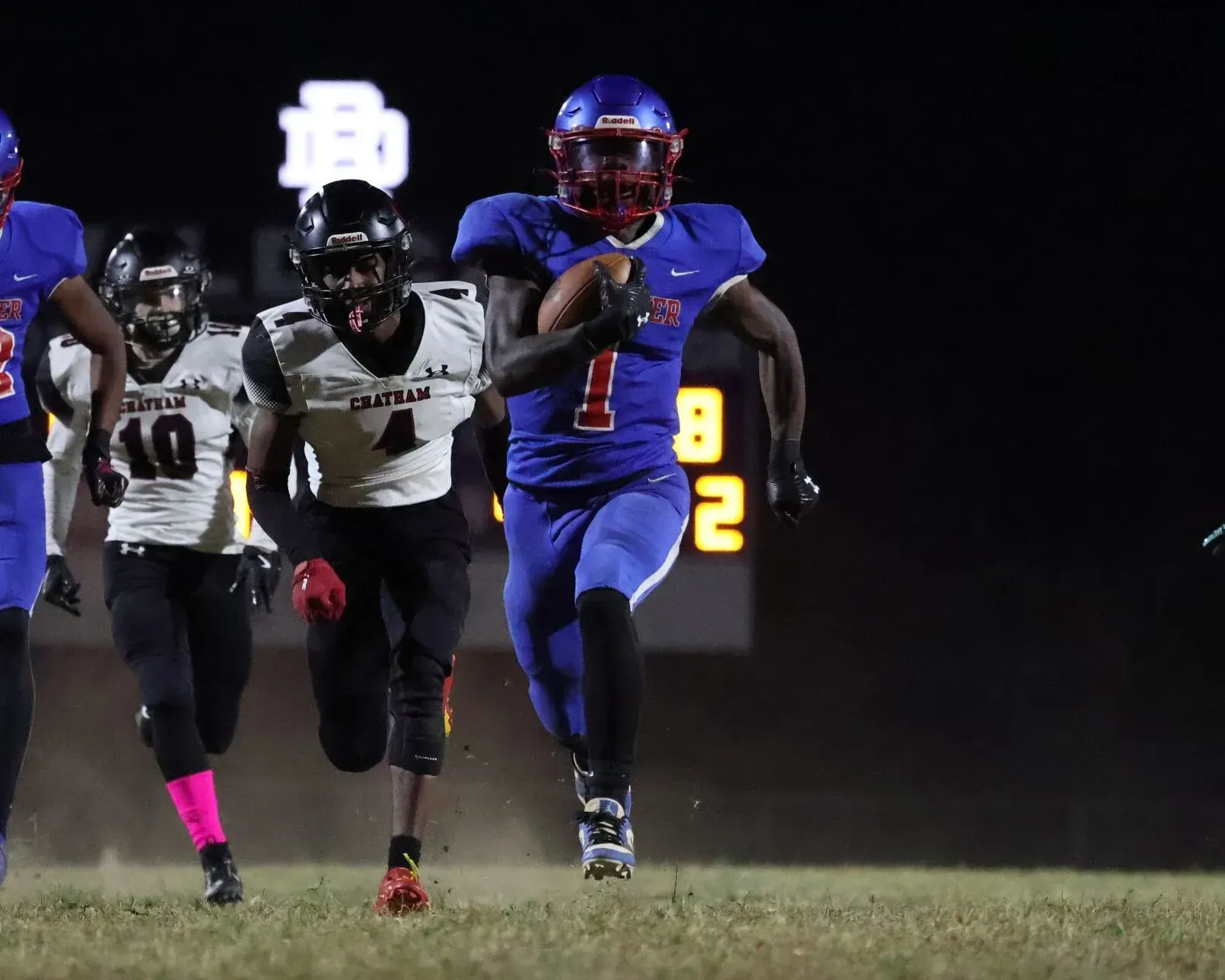 A football player in blue uniform runs with the ball while pursued by two defenders in white on a field at night.