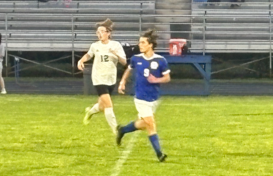 Two soccer players in blue and white jerseys running across a grass field toward the bleachers.