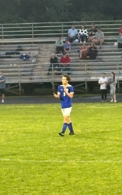 A soccer player in a blue jersey and white shorts walking on a grass field in front of stadium bleachers.