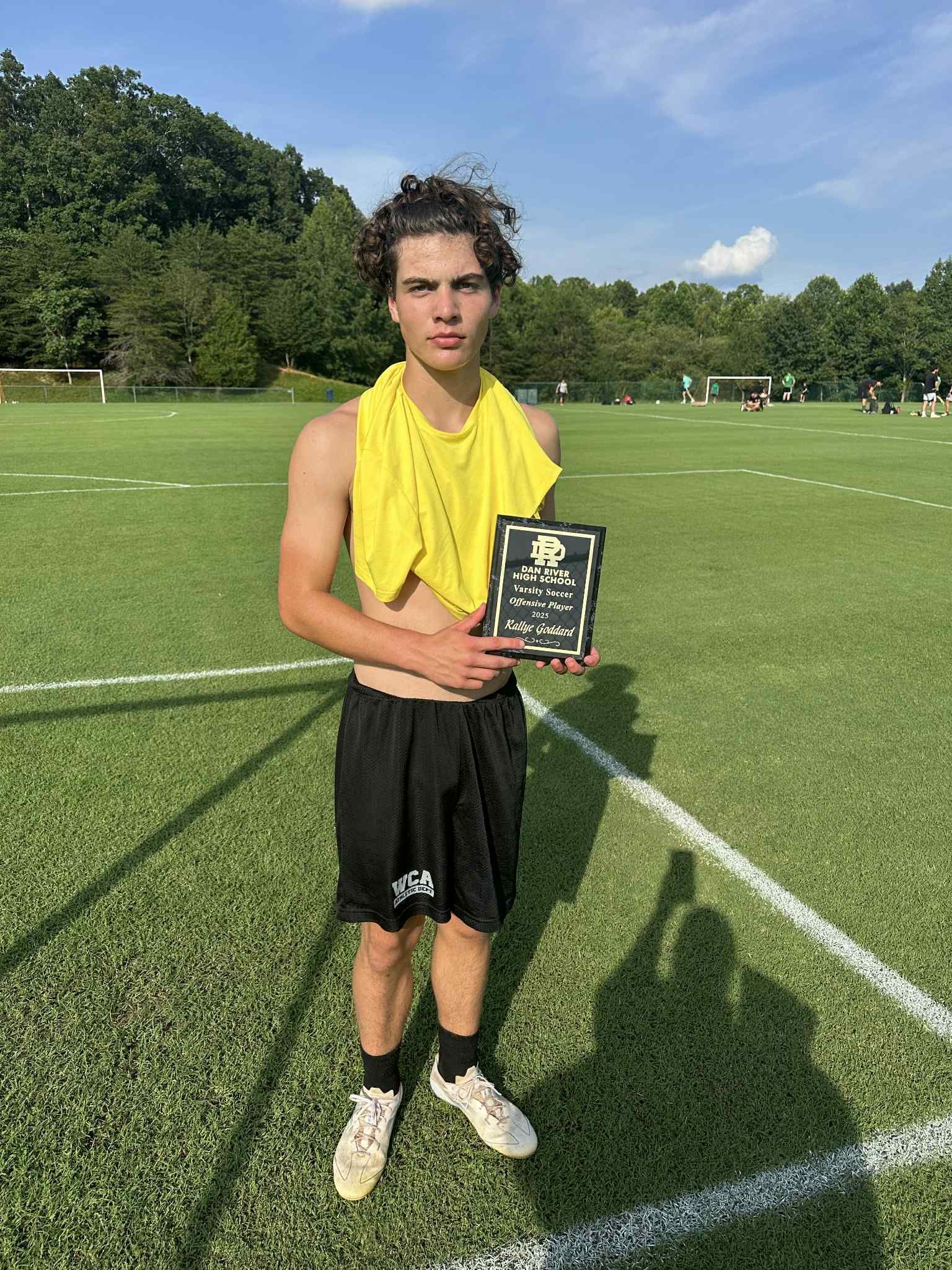 A person stands on a grass field holding a small award sign, wearing a yellow shirt draped over their shoulders and shorts.