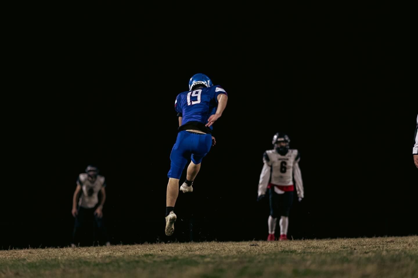 A football player in a blue uniform leaps through the air on a dark field, with two other players in the background.